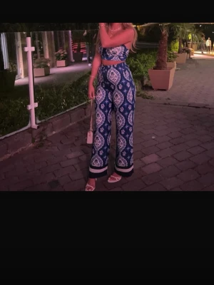 Blått mönstrat set med vida byxor - Säljer ett snyggt tvådelat set med bandeau-topp och vida byxor i blått och vitt paisleymönster. Setet har elastisk topp och byxor med hög midja och breda ben. Perfekt för sommaren och ger en bohemisk vibe. Materialet känns lätt och luftigt.