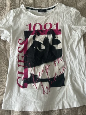 Vit t-shirt med tryck från Guess - Vit t-shirt från Guess med stort grafiskt tryck i svart, rosa och vitt framtill. Texten 'GUESS 1981' och 'Keep on dreamin’' i rosa ger en cool vibe. Klassisk rund hals och korta ärmar. Perfekt för en trendig och avslappnad stil.