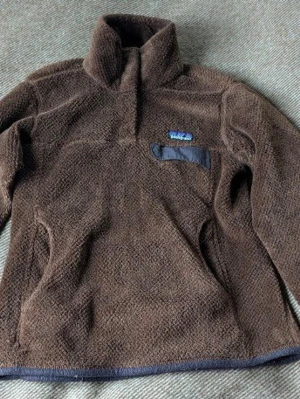 Brun fleece-tröja från Patagonia - Mysig brun fleece-tröja från Patagonia med halv dragkedja och hög krage. Tröjan har en liten bröstficka med dragkedja och broderad logga. Perfekt för kyliga dagar och enkel att slänga på sig över en t-shirt. Storlek small. Använt skick med lite noppor men inget stort som syns. Pris kan absolut diskuteras