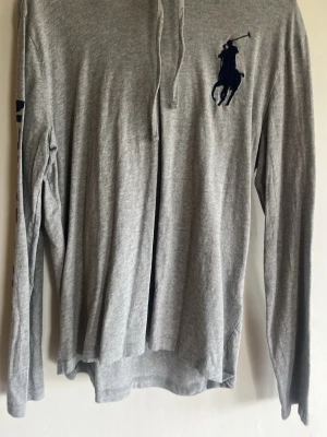 Grå långärmad topp från Polo Ralph Lauren - Snygg grå långärmad tunn luvtröja från Polo Ralph Lauren med klassisk mörkblå broderad logga på bröstet. Tillverkad i mjuk bomull och har en enkel knappslå framtill. Perfekt till jeans för en clean och sportig look. Även tryck på ena ärmen och bak. Se bilder.