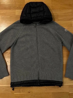 Grå cardiganjacka från Moncler - Grå stickad cardiganjacka från Moncler med svart quiltad huva och nederkant till ett galet bra pris pga flytt. Jackan har hel dragkedja framtill, ribbstickade muddar och Moncler-logga på ärmen. Perfekt för lager-på-lager och snygg till streetwear. Sitter som M. Pris kan diskuteras minimalt vid snabb affär. Passa på och göra en fantastisk affär.