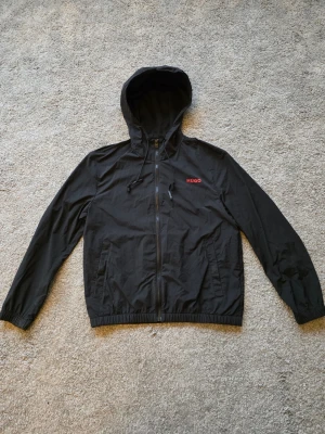 Svart vindjacka från HUGO med huva - Svart vindjacka från HUGO med dragkedja framtill, justerbar huva med snören och två sidofickor. Jackan har röd HUGO-logga på bröstet och elastiska muddar vid ärmslut och midja. Perfekt för blåsiga dagar och enkel att matcha till streetwear.