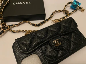  svart quiltat mobilskal med kedja - Stilrent svart mobilskal  i quiltat läder med guldfärgad kedja. Skalets design har en liten ficka framtill och kedjan är flätad med läderdetaljer. Perfekt accessoar för dig som vill ha både lyx och funktion.