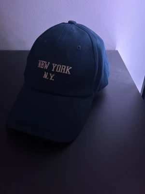 Mörkblå keps med New York-brodyr - Snygg mörkblå keps med vit brodyr där det står 'NEW YORK N.Y.' framtill. Klassisk böjd skärm och justerbart spänne bak. Tillverkad i bomullstyg för en bekväm känsla. Perfekt accessoar för en avslappnad streetwear-look. Helt ny