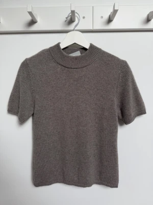 Tröja i kashmir  - Supersoft brun/taupe färgad stickad topp från H&M Premium Selection i 100% kashmir. Modellen har korta ärmar, ribbad hög hals och en clean, minimalistisk look. Perfekt till jeans eller kjol för en chill men ändå snygg vibe. Tröjan kan tvättas i 30 grader ullprogram/handtvätt. Storlek S, längd fram från halskanten 53 cm, bredd under ärmen 43 cm, längd ärm 23 cm.  Nypris 1350 kr.