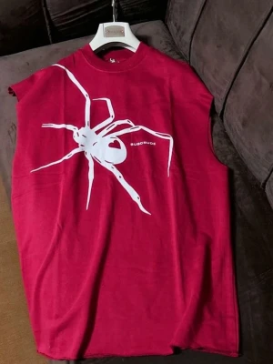 Röd ärmlös t-shirt med spindeltryck - Cool röd ärmlös t-shirt från Subcrude med stor vit spindelgrafik på framsidan. Oversized passform och rund halsringning. Perfekt för dig som vill sticka ut med en unik och edgy stil.