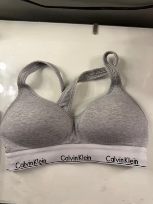 Grå sport-bh från Calvin Klein - Grå sport-bh från Calvin Klein med vadderade kupor och bred resår med logga. Bh:n har korsade axelband och är tillverkad i mjuk bomullsblandning för extra komfort. Säljer för är för liten för mig❤️