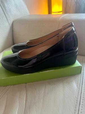 Svarta lackade ballerinaskor med kilklack - Snygga svarta ballerinaskor i lackat material med diskret kilklack. Skorna har rund tå och en klassisk, stilren design som passar till många outfits. Insidan är ljusbeige och skon har en mjuk innersula för extra komfort.i skinn. 