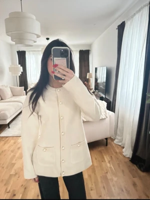 Krämvit kofta med pärlknappar - Snygg krämvit kofta med pärlknappar och två fickor framtill. Koftan har rund halsringning och ribbade detaljer vid knäppningen. Perfekt för dig som gillar en stilren och elegant look.