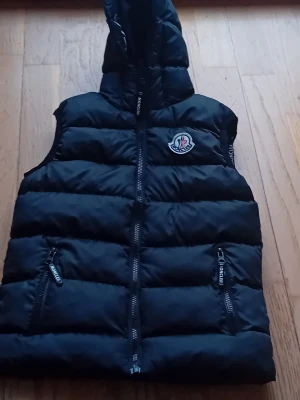 Svart dunväst från Moncler med huva - Snygg svart dunväst från Moncler med huva och dragkedja framtill. Västen har två dragkedjeförsedda fickor och det klassiska Moncler-logotypen på bröstet. Perfekt för lager-på-lager och streetstyle. Materialet är glansigt och quiltat för extra värme. Priset kan diskuteras.