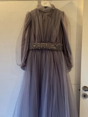 Lång lila tyllklänning med paljettbälte - Elegant långklänning i lila tyll med puffiga långärmar och hög hals. 