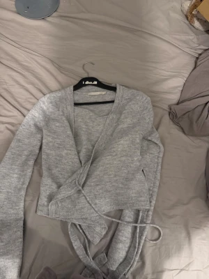 Grå omlottkofta från Lager 157 - Snygg grå omlottkofta från lager 157 med långa ärmar och knytband i midjan. Koftan är stickad och har en mjuk, skön känsla. Perfekt för lager-på-lager och enkel att matcha med olika outfits.