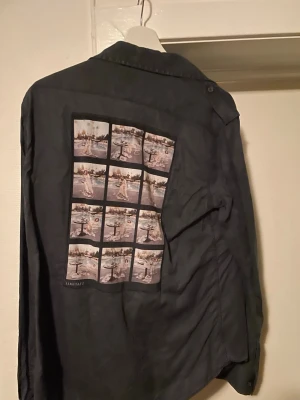 Svart Limitato Skjorta L - Cool svart skjorta från Limitato med knappar framtill och två bröstfickor. På ryggen finns ett stort fotoprint med flera bilder i rutmönster, vilket ger skjortan en unik och kreativ look. Perfekt för dig som gillar streetstyle och vill sticka ut.