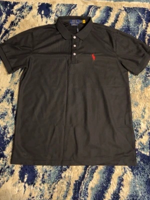 Svart pikétröja från Polo Ralph Lauren - Säljer en svart pikétröja från Polo Ralph Lauren med klassisk krage och knappar framtill. Tröjan har korta ärmar och ett rött broderat logomärke på bröstet. Perfekt för dig som gillar stilrena plagg med ikonisk design.