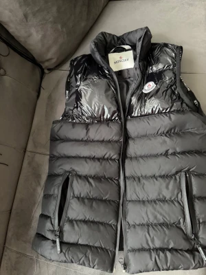 Svart dunväst från Moncler - Snygg svart dunväst från Moncler med glansig ovandel och klassisk Moncler-logga på bröstet. Västen har dragkedjor på fickorna och en cool serietidningsetikett på insidan. Perfekt för lager-på-lager och streetstyle.