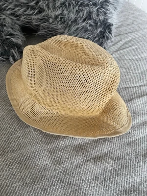 Beige stråhatt med klassisk form - Snygg beige stråhatt med klassisk fedora-form och bredare brätte. Hatten är tillverkad i flätad strå och har en luftig, öppen struktur som passar perfekt för soliga dagar. Enkel och stilren design utan synligt märke.