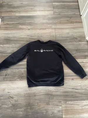 Mörkblå Sail Racing sweatshirt - Mörkblå sweatshirt från Sail Racing med vit logga tryckt på bröstet. Tröjan har rund halsringning, långa ärmar och ribbade muddar vid ärmslut och nederkant. Perfekt för en avslappnad och sportig stil.