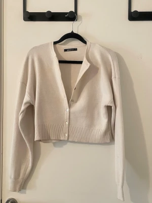 Beige cropped kofta från Gina Tricot - Cropped kofta i vit från Gina Tricot med långa ärmar och ribbade muddar. Koftan har v-ringning och knäpps med fyra vita knappar framtill. Perfekt att slänga över en topp för en chill look.