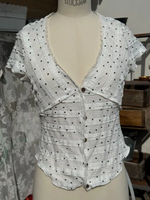 Vit prickig blus med knappar - Söt vit blus med prickar och korta ärmar. Djup V-ringning och dekorativa knappar framtill. Tillverkad i ett lätt, strukturerat tyg med vågig nederkant och spetsdetaljer vid halsen. Figurnära passform som ger en feminin siluett.