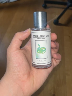 Goldhaber 27.0 parfym från One Bold Chemist - Goldhaber 27.0 är en parfym i en genomskinlig glasflaska med silverfärgat lock. Flaskan har en modern etikett med en grön orm och svart text. Parfymen har en stilren och minimalistisk design som passar perfekt på hyllan.