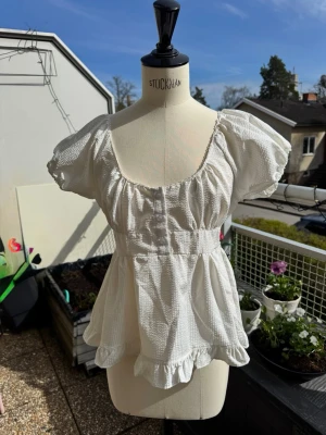 Vit blus med volang och puffärm - Supersöt vit blus med puffiga korta ärmar, markerad midja och volangkant nertill. Blusen har en lätt strukturerad bomullskänsla och en feminin, luftig passform. Perfekt för en fräsch och somrig look.