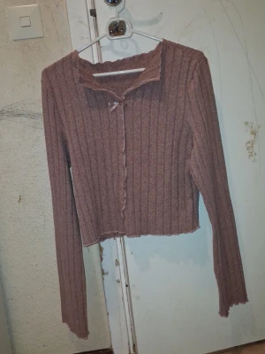 Ribbad brun kofta med volangkant (För kvinnor) - Säljer en ribbad brun kofta med volangkant längs krage, ärmslut och nederkant. Koftan har långa ärmar och en söt liten knytning framtill vid halsen. Perfekt för dig som gillar en trendig och feminin look.