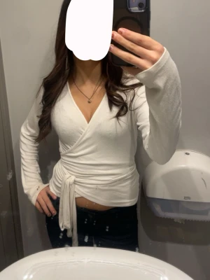 Tröja med  knytband - Säljer en vit omlottopp med knytband i midjan och v-ringad hals. Toppen har långärmade ärmar och är croppad, vilket ger en trendig look. Perfekt att matcha med jeans eller kjol för en snygg outfit.