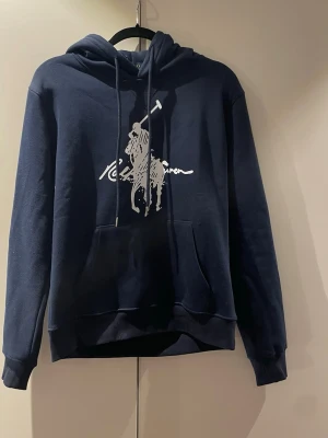 Mörkblå hoodie Polo Ralph Lauren - Helt ny Polo Ralph Lauren hoodie i marinblå färg med stor vit klassisk Polo-logga på bröstet. Storlek M, aldrig använd med alla taggar kvar. Tjock och skön bomullsfleece, bra passform med ficka och huva. Perfekt vardagsplagg!