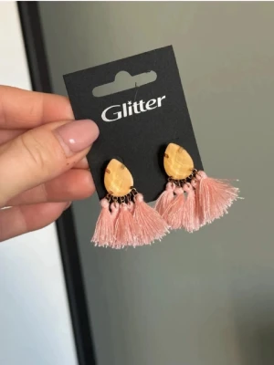 Glitter örhängen med rosa tofsar - Snygga örhängen från Glitter med droppformad guldfärgad sten och flera ljusrosa tofsar i tyg. Perfekta för att ge din look en trendig och lekfull touch. Fästs med stift och har en unik kombination av färg och form.