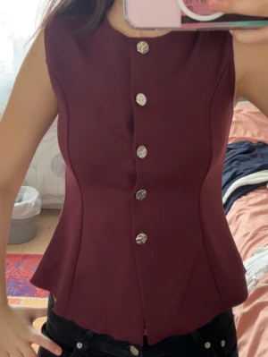 Vinröd väst med silvriga knappar - Snygg vinröd väst med figurnära passform och dekorativa silvriga knappar framtill. Västen har en stilren, ärmlös design och lätt utsvängd nederkant. Perfekt att styla över en skjorta eller t-shirt för en trendig look.