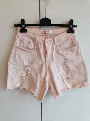Ljusrosa jeansshorts från Paris - Unika och snygga ljusrosa jeansshorts som köptes i en liten boutique i Paris för flera år sedan. Shortsens kanter är råa och fransiga med slitna detaljer framtill. Klassisk femficksmodell med knapp och dragkedja. I bra skick men fått lite diskreta gula missfärgningar från solkräm på insidan av midjan och längst ner på benen (svårt att fånga på bild, mycket diskret att knappt syns). Väldigt snygg passform och har varit älskade. 