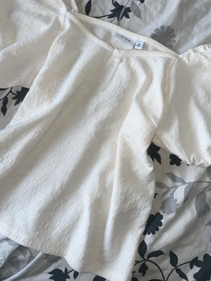 Vit blus med puffärm från NA-KD - Superfin vit blus från NA-KD med puffiga korta ärmar och v-ringning. Blusen har en lätt strukturerad yta och en liten knappdetalj framtill. Perfekt att styla med jeans eller kjol för en trendig look.