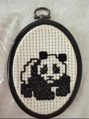 Broderad panda i svart ram - En oval broderad tavla med en svartvit panda i pixelstil. Motivet är broderat med svart garn på vitt tyg och ramen är svart med en metallögla för upphängning. Perfekt som unik dekoration eller present till någon som gillar handgjorda detaljer.