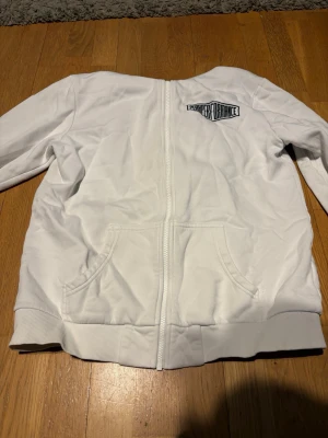 Vit hoodie med dragkedja - Snygg vit hoodie med dragkedja framtill och två fickor. Märket LeetStreet Ordnance är tryckt på bröstet. Hoodien har ribbade muddar och en klassisk huva. Perfekt för en chill och avslappnad stil. Sitter som en s/xs