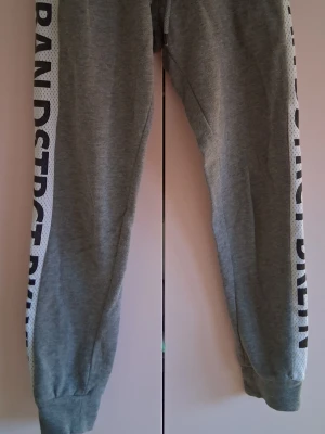 Grå mjukisbyxor med text på sidan - Säljer ett par grå mjukisbyxor från Urban District med breda vita ränder och svart text längs sidorna. Byxorna har resår i bensluten och är perfekta för chill dagar eller träning. Tillverkade i mjukt bomullsmaterial.