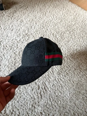 Svart keps med GG-mönster säljs för 200 kronor  - Snygg svart keps med klassiskt GG-monogram över hela kepsen och en grön-röd rand på sidan. Justerbar rem baktill och detaljer i bomullscanvas. Perfekt för dig som vill ha en exklusiv och trendig accessoar.