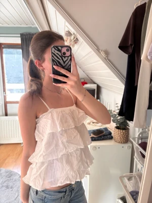 Vit volangig linnetopp - Säljer en söt vit linnetopp med tunna axelband och flera lager volanger. Toppen är luftig och har en avslappnad passform, perfekt för varma dagar. Passar till jeans eller kjol för en trendig sommarlook.