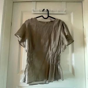 Beige blus med rynkade detaljer - Säljer en beige blus med korta ärmar och rynkade detaljer i midjan. Blusen har en lös passform och är tillverkad i ett lätt, skrynkligt material som ger en avslappnad look. Perfekt att styla med jeans eller kjol.