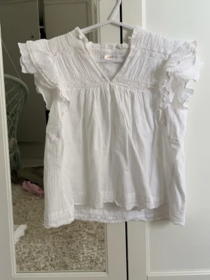 Vit volangblus med spetsdetaljer - Söt vit blus med volangprydda ärmar och v-ringning. Blusen har fina spetsdetaljer och lätt rynk vid axlar och bröst. Tillverkad i ett tunt, luftigt tyg som känns som bomull. Perfekt för en fräsch och somrig look.