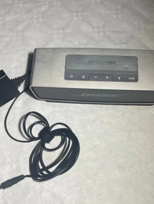 Bose SoundLink Mini trådlös högtalare - Snygg och kompakt trådlös högtalare från Bose, modell SoundLink Mini. Högtalaren är silverfärgad med en robust aluminiumkropp och har tydliga knappar för volym, bluetooth och AUX. Perfekt för att streama musik via bluetooth eller ansluta med kabel.