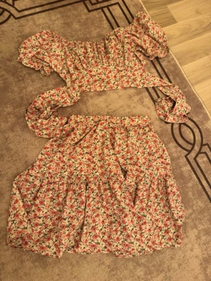 Blommigt tvådelat set med puffärm - Säljer ett sött tvådelat set med blommigt mönster i rosa, vitt och grönt. Toppen har puffärmar och knytband, kjolen är kort och har resår i midjan. Setet är gjort i ett lätt, luftigt tyg som känns perfekt för varma dagar.