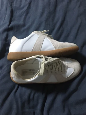  sneakers -  sneakers i vitt och beige läder och mocka med klassisk gummisula. Låga sneakers med snörning och stilren design, perfekta för en clean och trendig look. Märkning på insidan av skon.