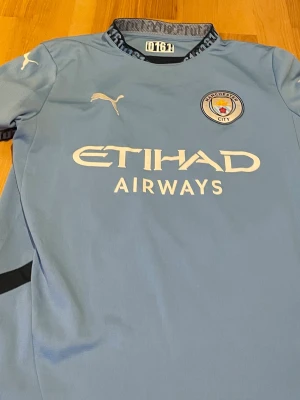 Manchester City matchtröja Foden 47 - Snygg ljusblå Manchester City matchtröja från Puma med Etihad Airways-tryck på bröstet och Foden 47 på ryggen. Tröjan har klubbmärke på bröstet, korta ärmar och detaljer i svart och vitt. Perfekt för fotbollsfans som vill stötta sitt lag.