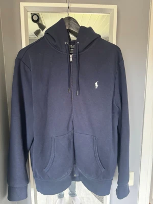  hoodie Ralph Lauren - Snygg mörkblå hoodie från Polo Ralph Lauren med dragkedja, huva med dragsko och två stora fickor framtill. Klassisk logga broderad på bröstet. Tillverkad i mjukt bomullsmaterial som är perfekt för chill dagar.