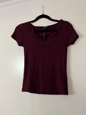 Vinröd topp med spetsdetalj - Snygg vinröd topp med v-ringning och spetsdetalj runt halsen. Kortärmad och figurnära passform, perfekt att styla med jeans eller kjol. Mjuk och skön känsla, enkel men ändå elegant look.