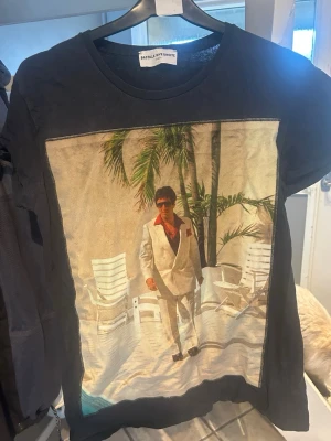 Svart t-shirt Bastille Le Droit - Svart t-shirt från Bastille Le Droit med stor färgglad print framtill som visar en person i vit kostym bland palmer och stolar. På ärmen finns en röd stjärna med palmtryck. Klassisk passform och rund hals. Perfekt för dig som gillar statement-plagg.