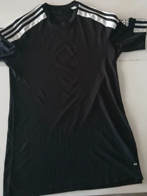 Svart Adidas sporttröja med ränder - Säljer en svart sporttröja från Adidas med klassiska vita ränder på axlarna och logga på ärmen. Tröjan har rund hals och är tillverkad i ett lätt, snabbtorkande material som passar perfekt till träning eller fotboll.