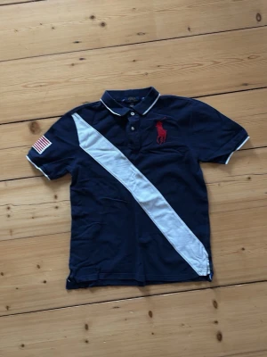Marinblå pikétröja från Polo Ralph Lauren - Marinblå pikétröja med vit diagonal rand, röd broderad logga på bröstet och amerikansk flagga på ärmen. Klassisk krage med knappar och korta ärmar. Perfekt för en sportig och stilren look.