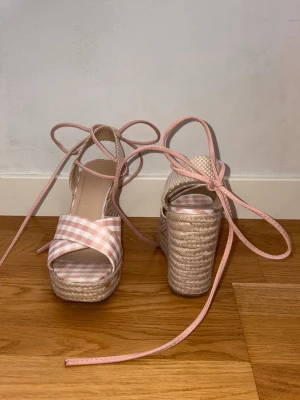 Rosa kilklackar med rosetter - Snygga platåsandaler med kilklack och snörning runt ankeln. Ovandelen har ett rosa och vitt rutigt mönster. Klacken är flätad för en somrig känsla. Perfekta för dig som vill sticka ut med en söt och trendig look.