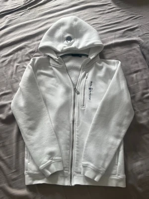 Vit zip-hoodie från Paul & Shark - Snygg vit hoodie med dragkedja från Paul & Shark. Hoodien har en broderad logga på bröstet och på huvan, samt en praktisk bröstficka med dragkedja. Mjuk insida och ribbade muddar. Perfekt för en clean och sportig look.